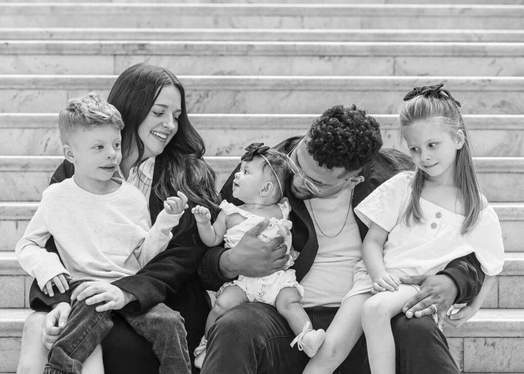 A mother, father, and their three young children sit on the marble steps and smile at each other for their family portraits at the Utah state capitol