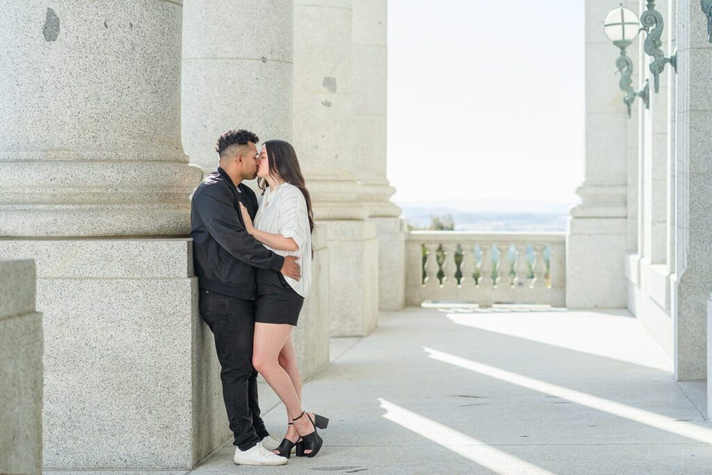 a man holds his girlfriend and kisses her as he leans against a granite column outside of the Utah state capitol rotunda