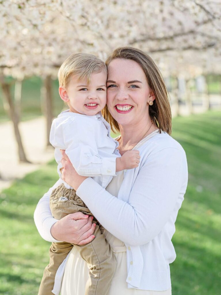 a mother wearing a white sweater and cream colored dress holds her toddler son with blossoming trees behind them in salt lake city