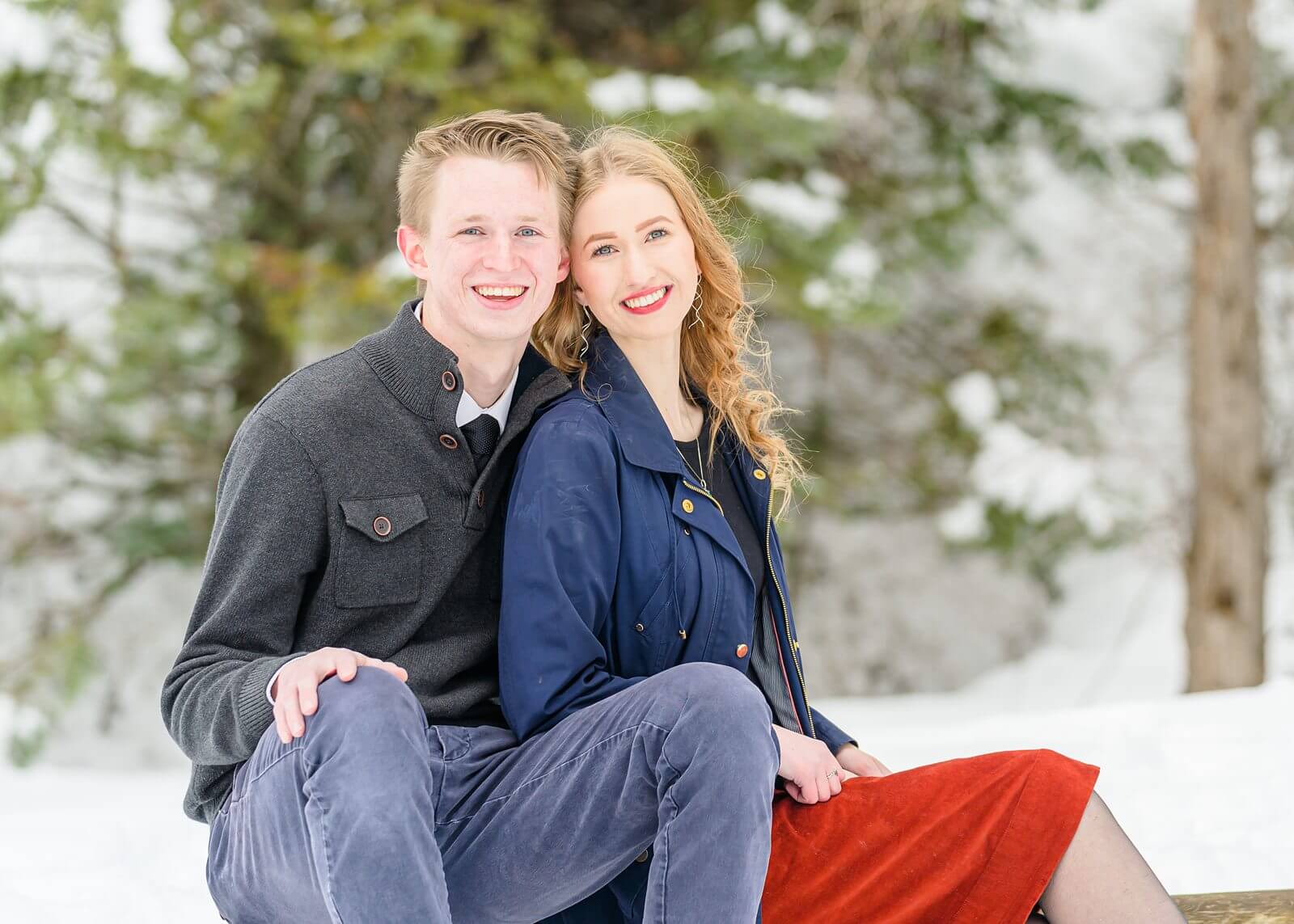 a man wearing a dark gray pullover and woman wearing a navy blue jacket at Tibble Fork Reservoir sit next to each other on a fence show for their winter engagement photos
