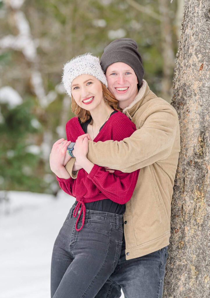 a man wearing a tan winter jacket leans against a tree and wraps his arms around his fiancee wearing a burgundy sweater and a white beanie for their engagement photos