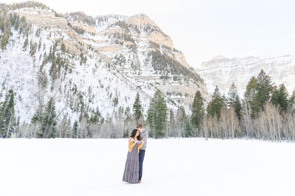 An engaged couple embrace in the snow with the stunning Provo Canyon mountains behind them