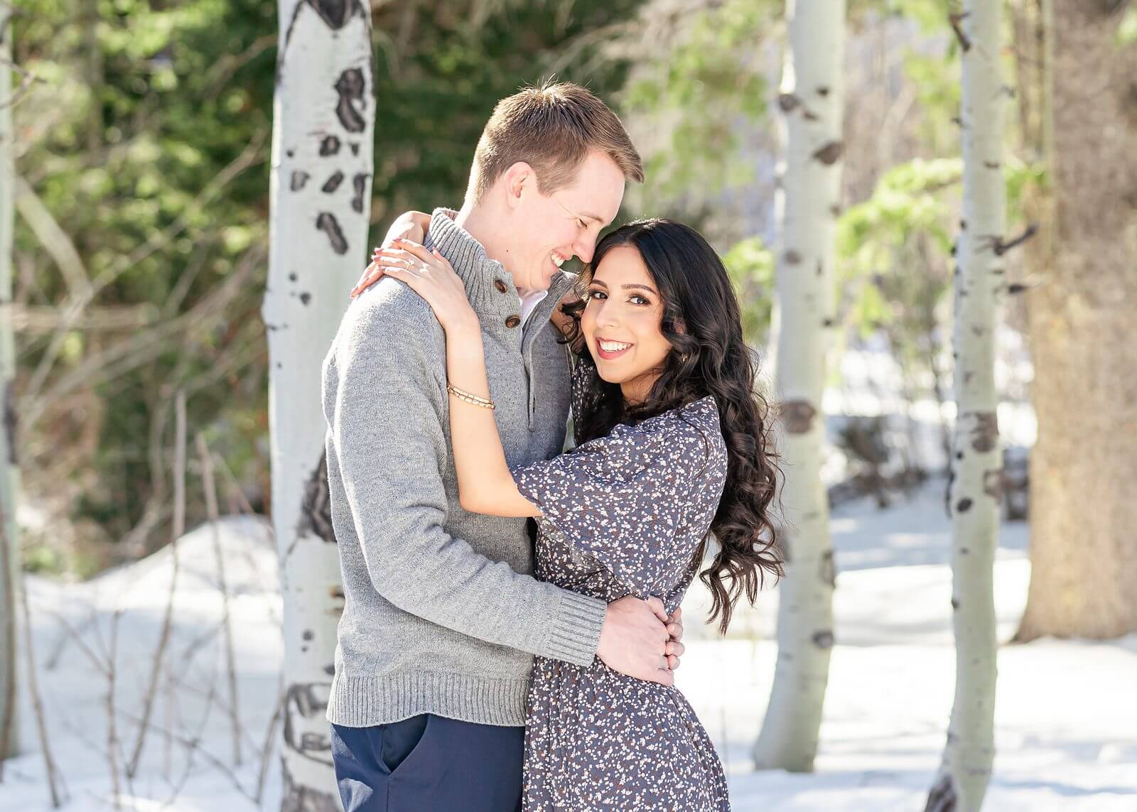 A man smiles at his fiancee as they stand in front of the trees for their winter engagement session in Provo Canyon