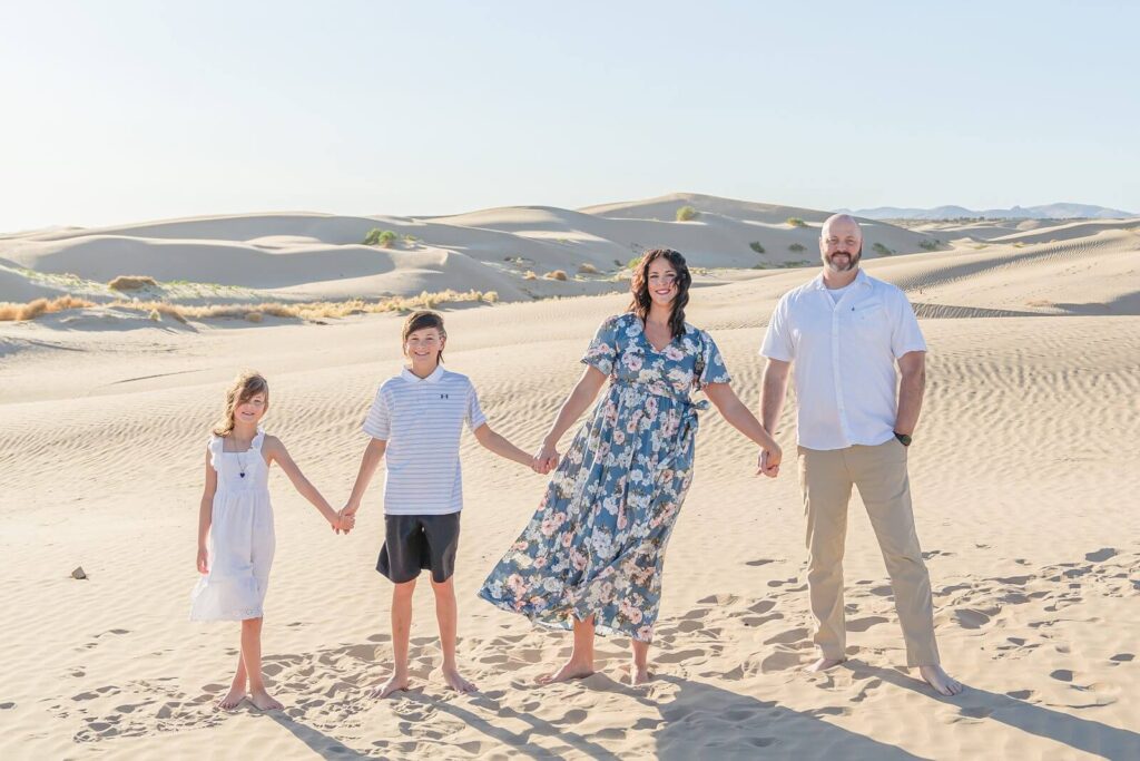 a mother wearing a blue floral dress and a father wearing a white short sleeve button-down shirt and khaki pants hold hands with their son and daughter wearing white and black as they hold hands while standing in the sand at little sahara in Nephi