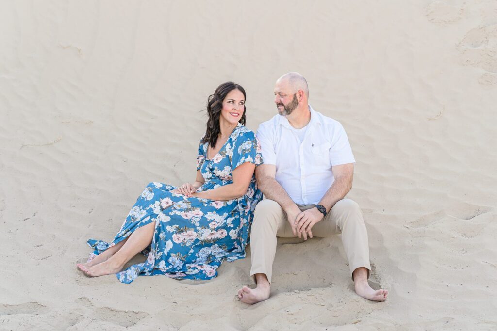 a woman wearing a long blue dress with white and pink flowers smiles at her husband wearing a white short sleeve button-down shirt and khaki pants as they sit in the sand in Nephi, Utah