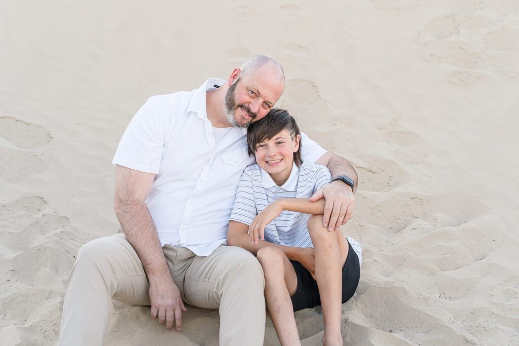 a father wearing a white short sleeve button-down shirt and khaki pants and his son wearing a white polo shirt and black shorts sit barefoot in the sand at Little Sahara in the summer