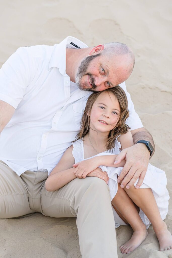 a father wearing a white short sleeve button-down shirt and khaki pants and his daughter wearing a white sundress sit barefoot in the sand at Little Sahara in the summer
