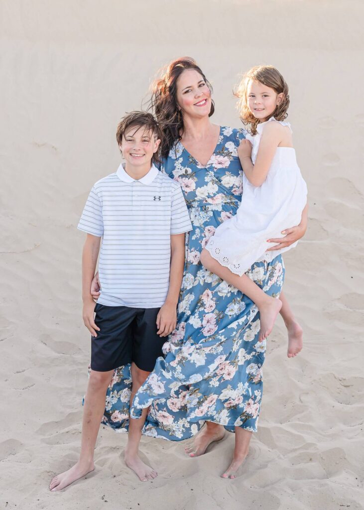 a wide shot of mother wearing a long blue dress with white and pink flowers and her daughter and son wearing white stand in the sand at Little Sahara in Nephi