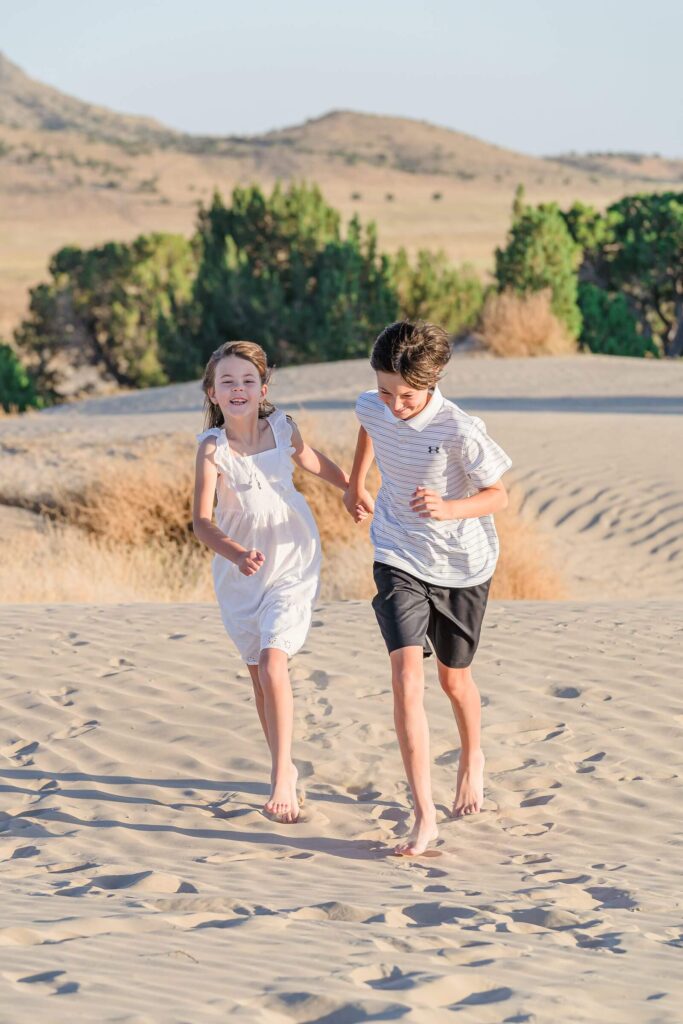 a girl wearing a white sundress and a boy wearing a white polo shirt and black shorts run in the sand at Little Sahara in Nephi, Utah