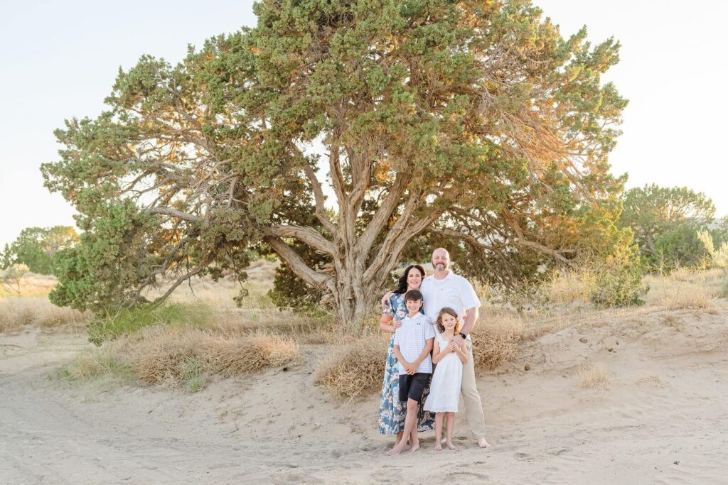 a mother, father, son, and daughter stand in front of a large juniper tree in the desert at sunset