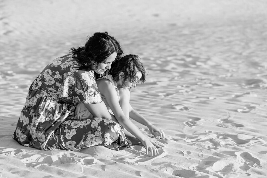 a black and white portrait of a mother and daughter playing in the sand at sunset in the summer