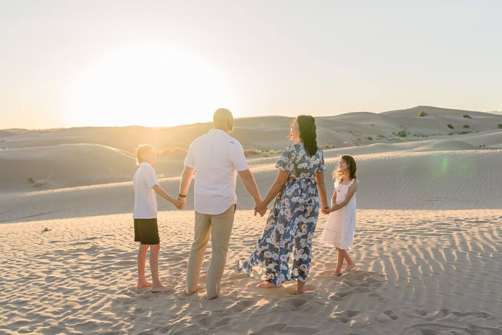a mother, father, and their son, and daughter hold hands while standing in the sand with the sun setting behind them at the little sahara sand dunes in Nephi in the summer