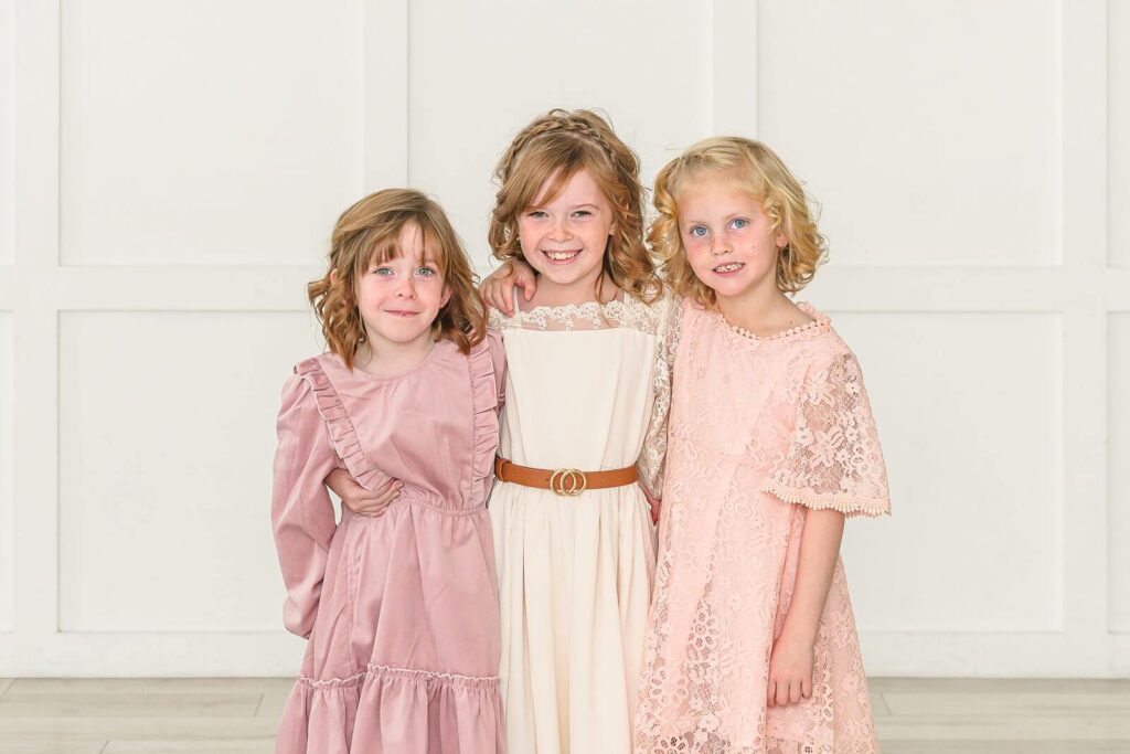 three young girls in pink, cream, and peach dresses smile during their family portrait session at The Blank Space Studios in American Fork
