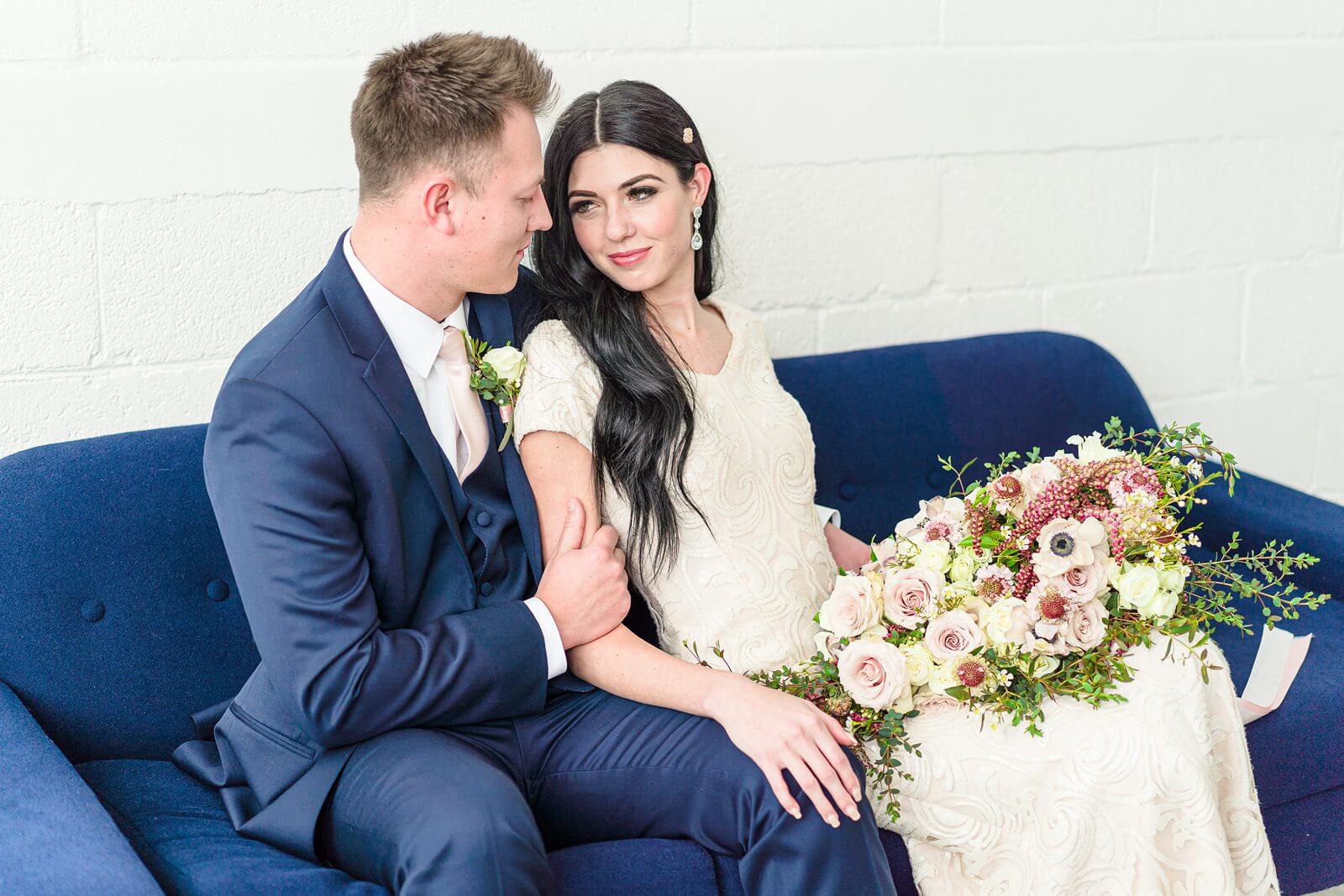 a groom wearing a navy blue and his bride wearing a cream colored short sleeved wedding dress sit on a navy colored couch at Studio Miesh in Salt Lake City