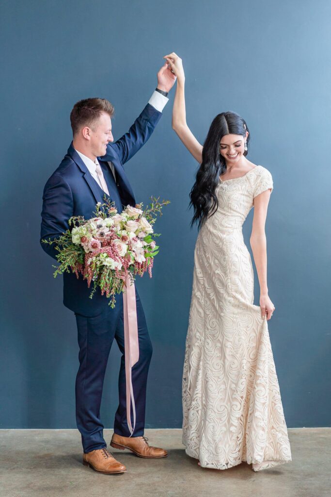 a groom wearing a navy blue suit and holding a large bouquet with pink and white roses twirls his bride wearing a cream colored wedding dress at Studio Miesh in Salt Lake City
