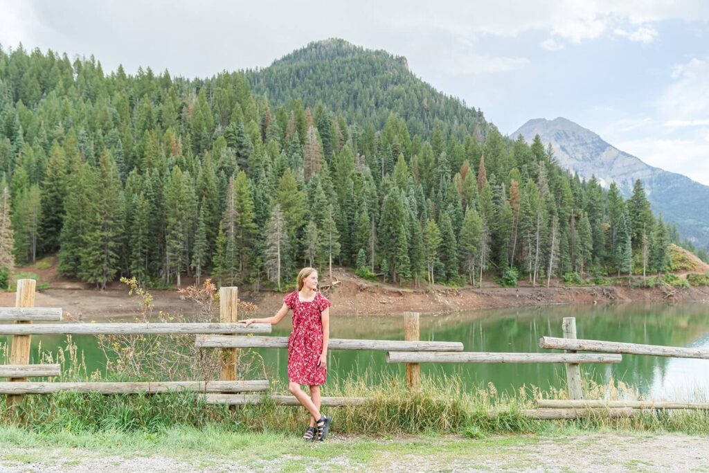 a high school senior girl wearing a red floral dress stands in front of a wooden fence in front of tibble fork reservoir in american fork