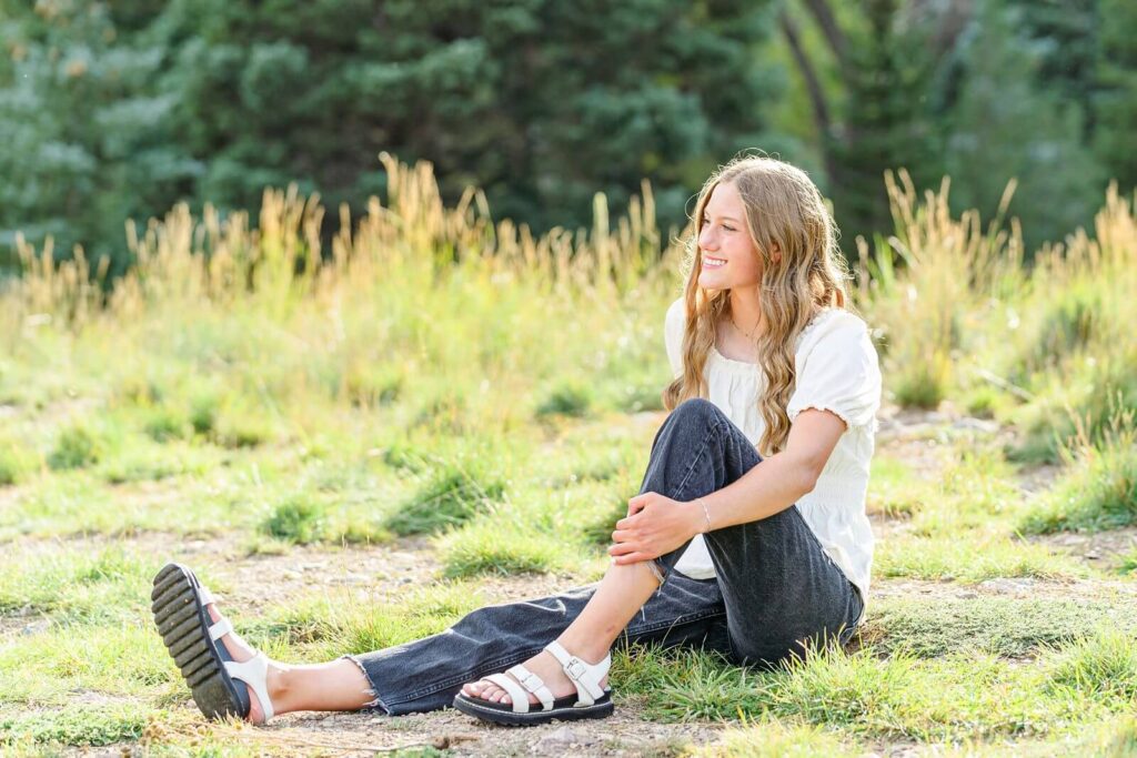 a senior girl from pleasant grove high school wearing a white short sleeved blouse and black jeans sits in the grass with the sun shining behind her in American Fork canyon