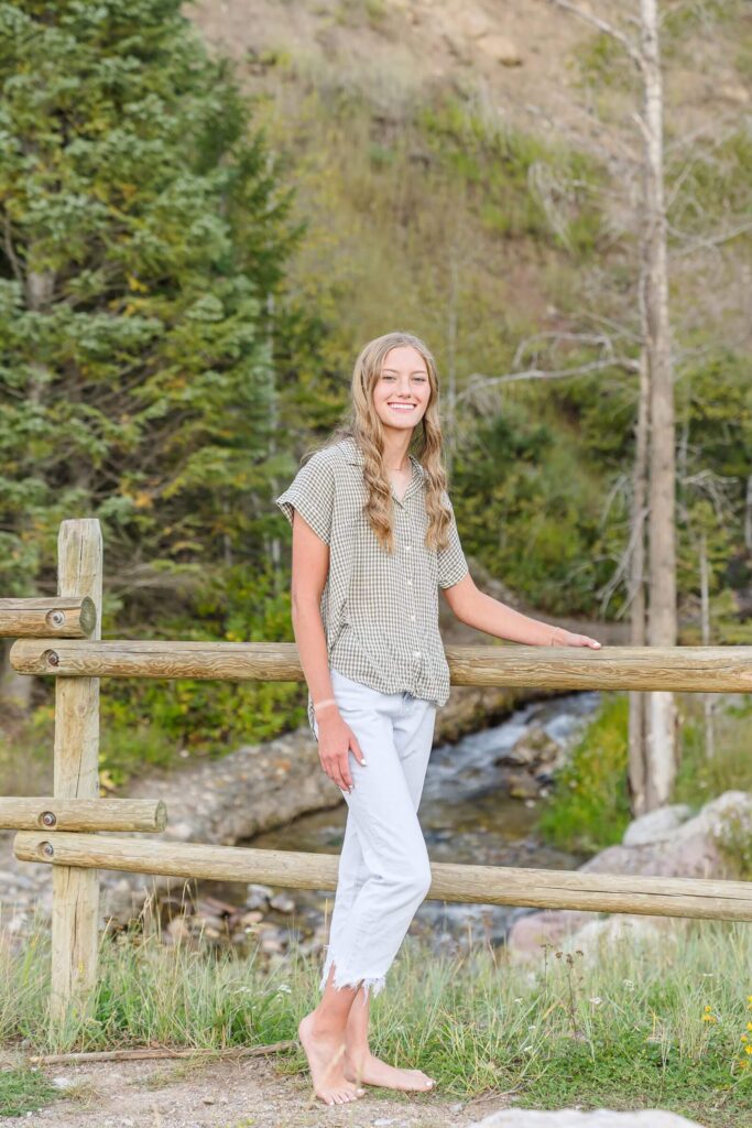 a high school senior girl wearing a green shirt, white jeans, and bare feet stands in front of a fence at Tibble Fork Reservoir in American Fork Canyon