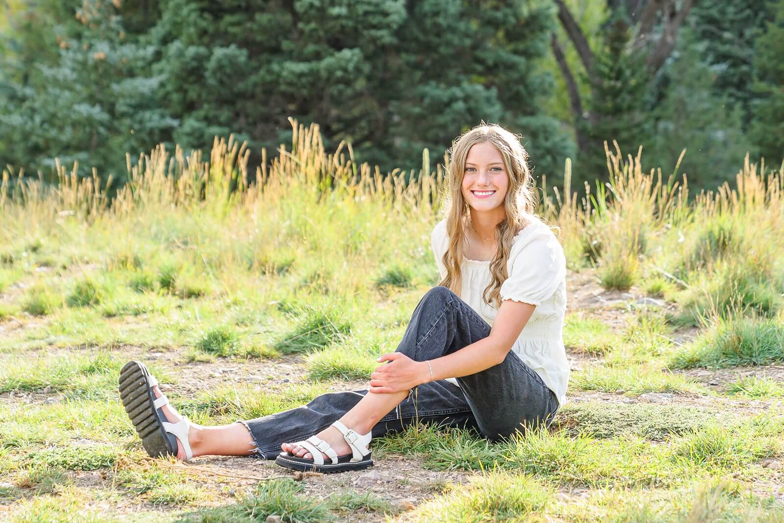 a girl wearing a white short sleeved blouse, black jeans, and white sandals sits in the grass for her senior photos at Tibble Fork Reservoir
