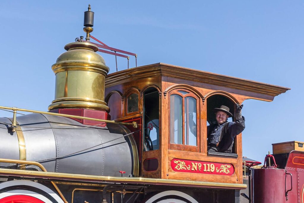 a train conductor leans out of a red and black steam engine and waves