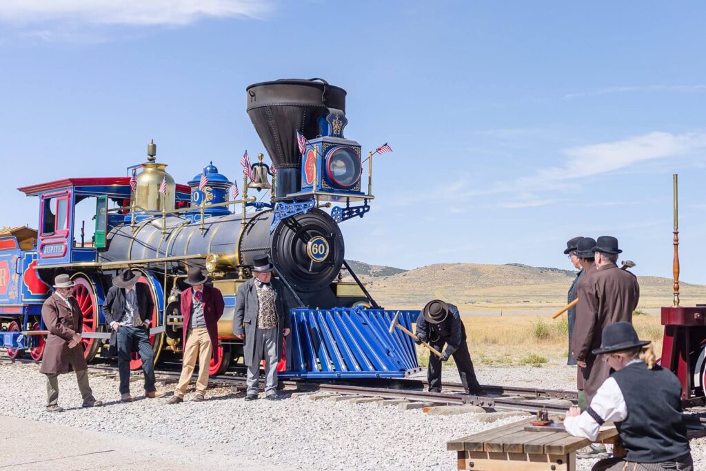 several men wearing clothes from the 1800's stand in front of a blue and black steam engine and reenact the famous driving of the golden spike at Promontory Summit in Utah