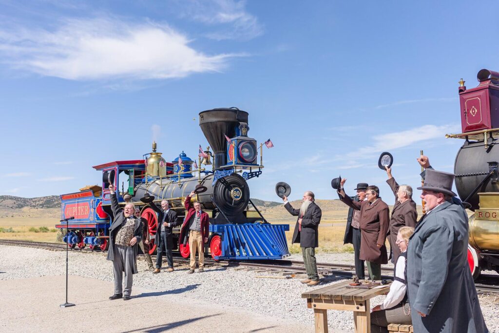 several men wearing clothes from the 1800's stand in front of a blue and black steam engine and cheer for the reenactment of the driving of the golden spike at Promontory Summit in Utah
