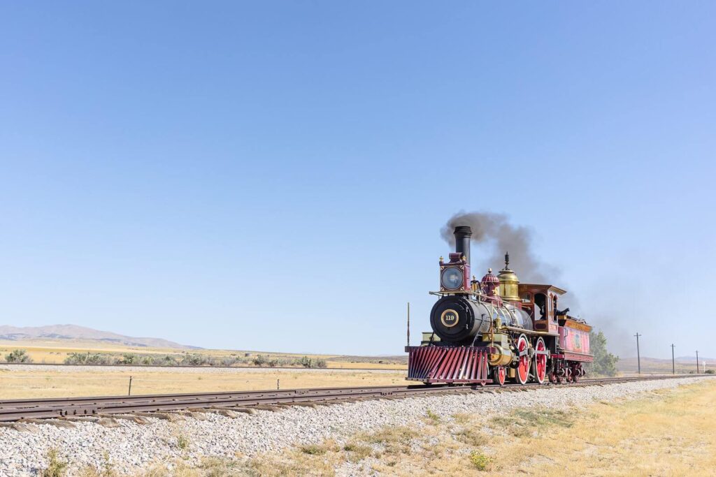 a beautiful black and red steam engine from the 1800's moves along the wooden tracks towards Golden Spike National Park
